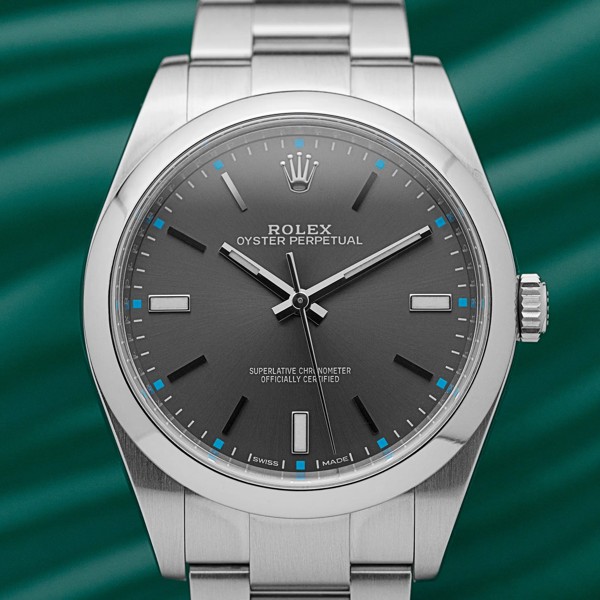 Front view of the Rolex Oyster Perpetual 39 (114300) with rhodium dial, blue square accents, and steel Oyster bracelet, on a green background.