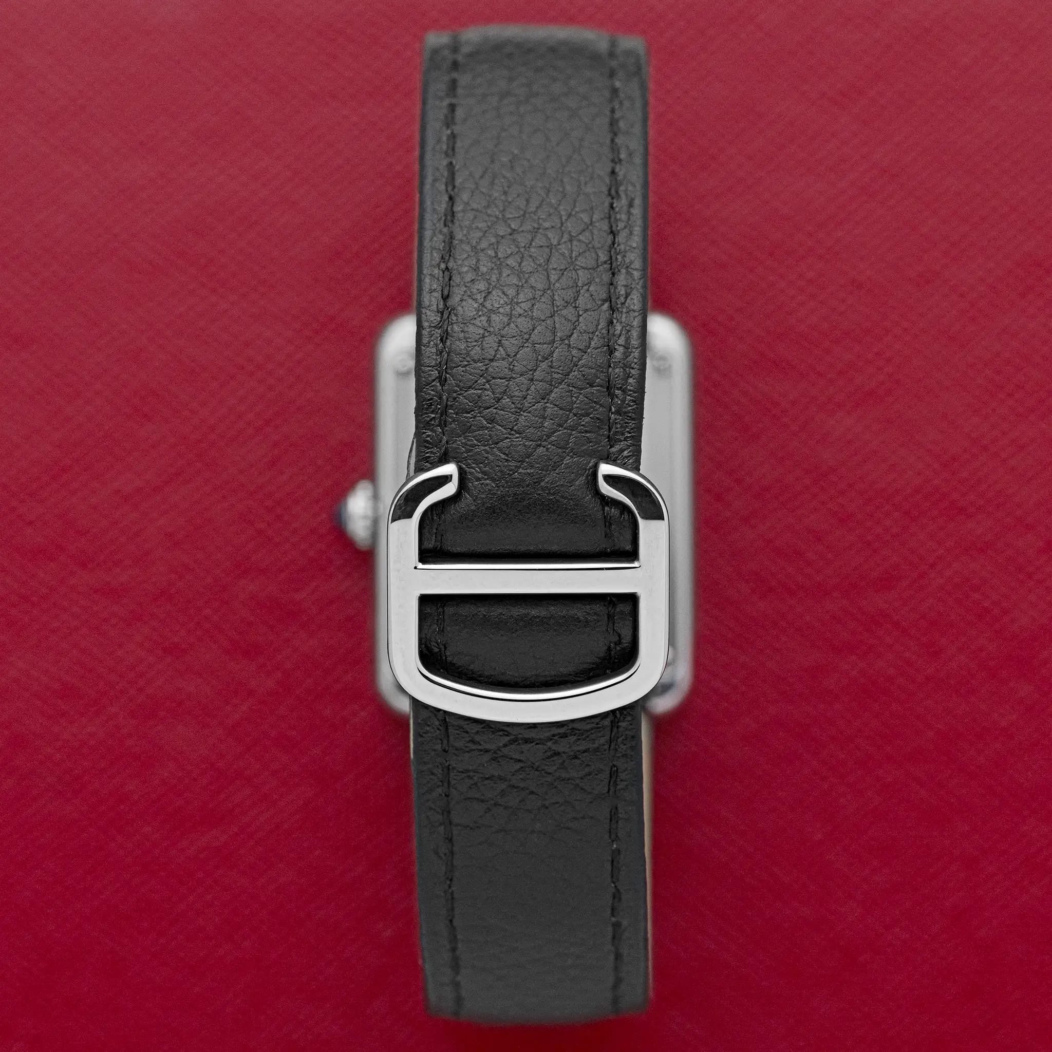 Close-up of the polished steel Cartier deployant clasp on the black leather strap, on a red background.