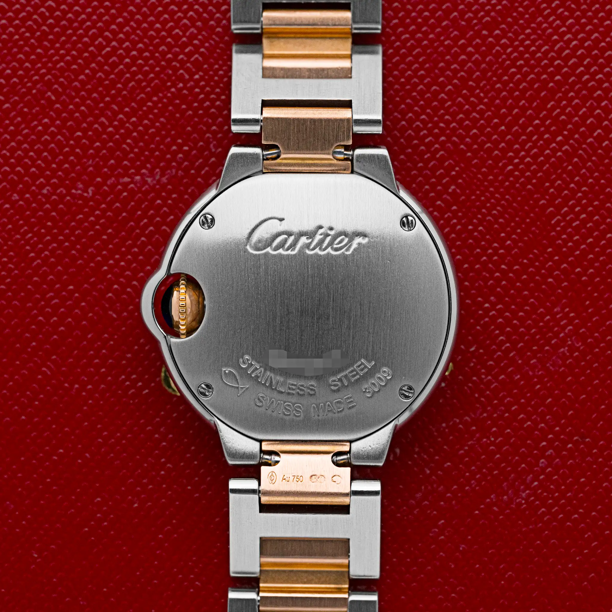 Steel case back of the Cartier Ballon Bleu 3009 engraved "Cartier, Stainless Steel, Swiss Made," with rose gold crown guard, on a red background.