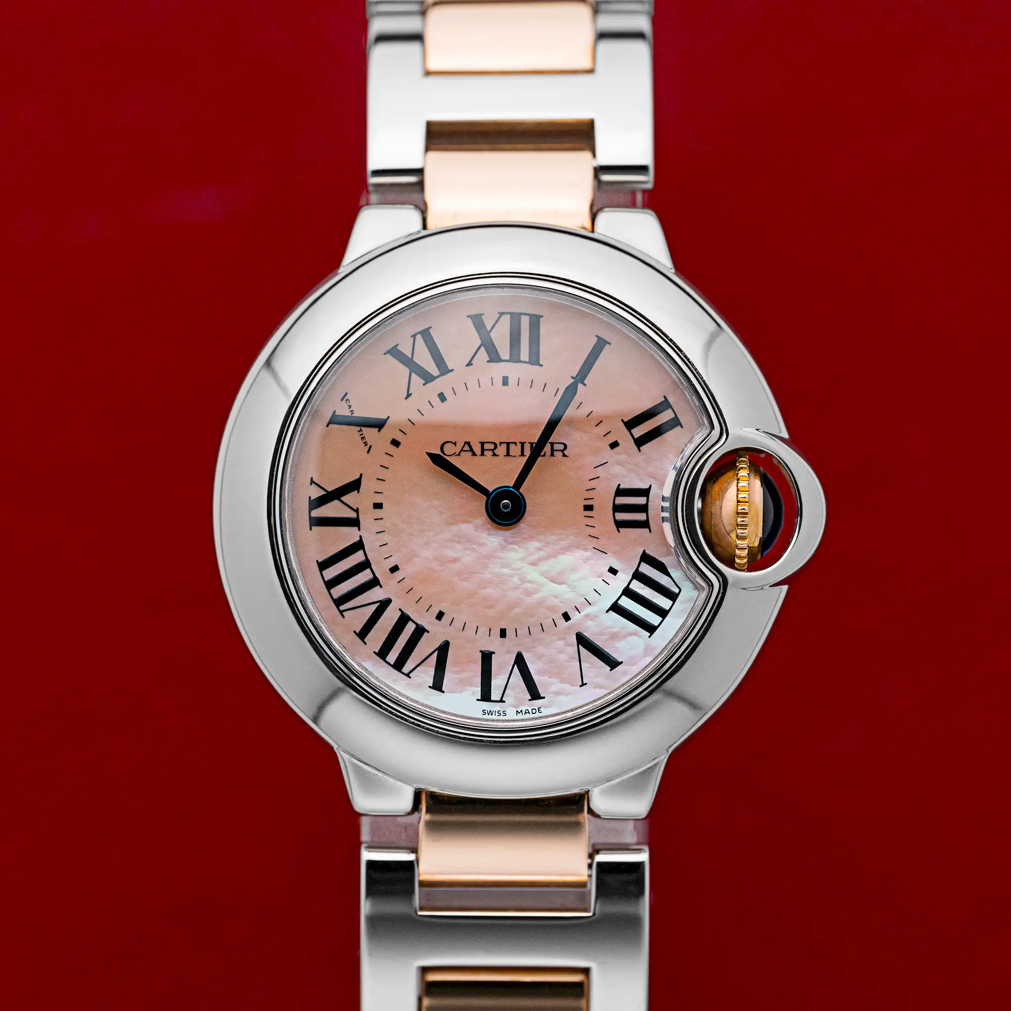 Front view of the Cartier Ballon Bleu 3009 with pink mother-of-pearl Roman dial, blued hands, and gold crown set with a blue cabochon, on a red background.